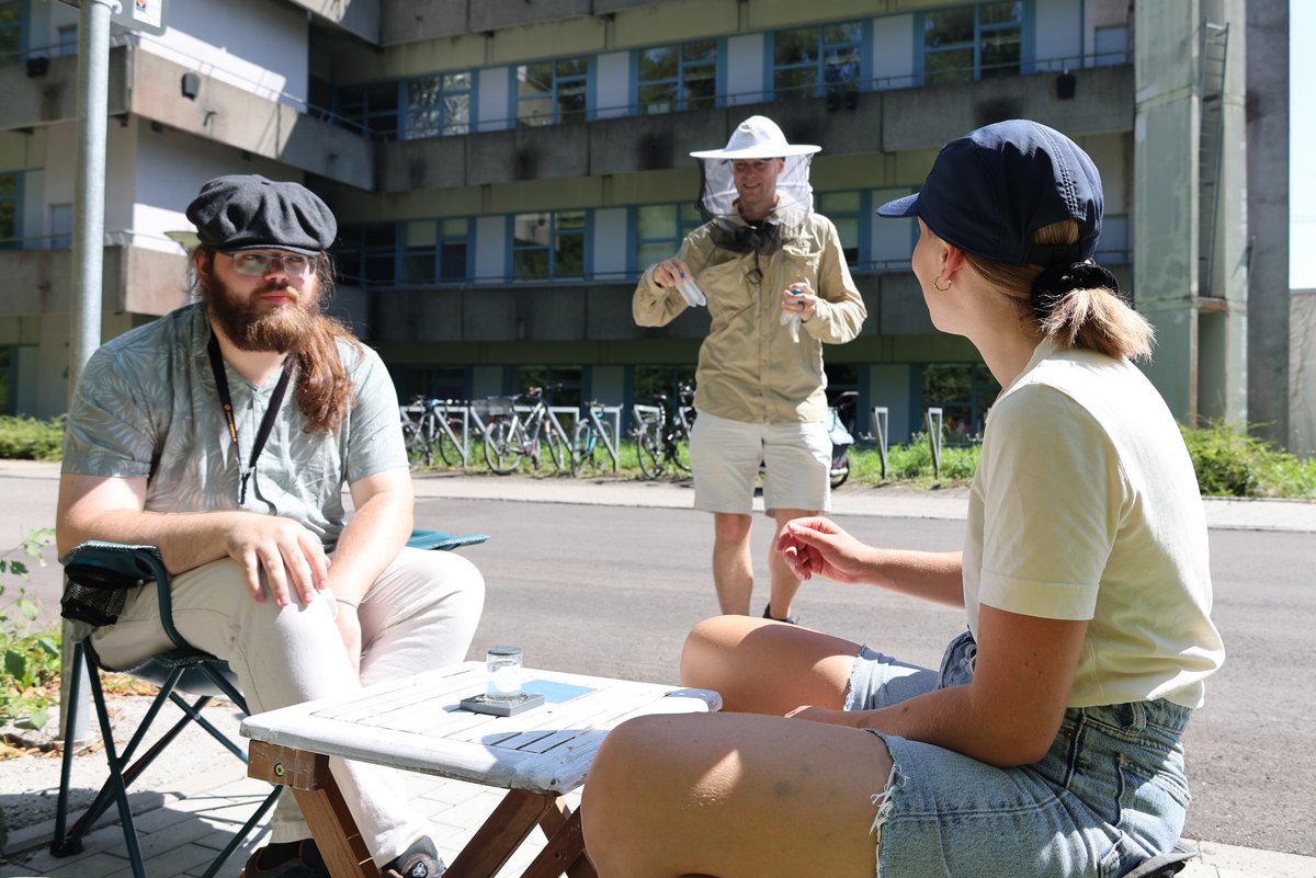 One person is wearing a beekeeper's helmet. Two people are sitting and waiting for him and the bees he carries.